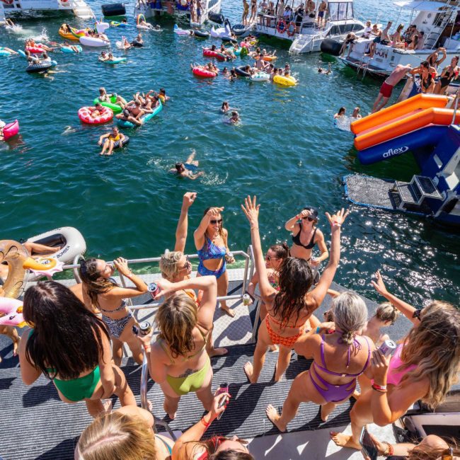 A group of people on a dock and in the water enjoying a sunny day; some are on inflatable rafts, others are on boats. The scene is lively with a luxury yacht hire Sydney adding an extra touch of elegance to the fun-filled day.