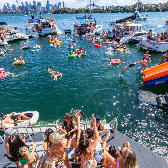 People enjoying a sunny day on a lake with boats and inflatables. Some are swimming, while others are on the deck of a private yacht charter in Sydney Harbour. The city skyline and bridge are visible in the background.