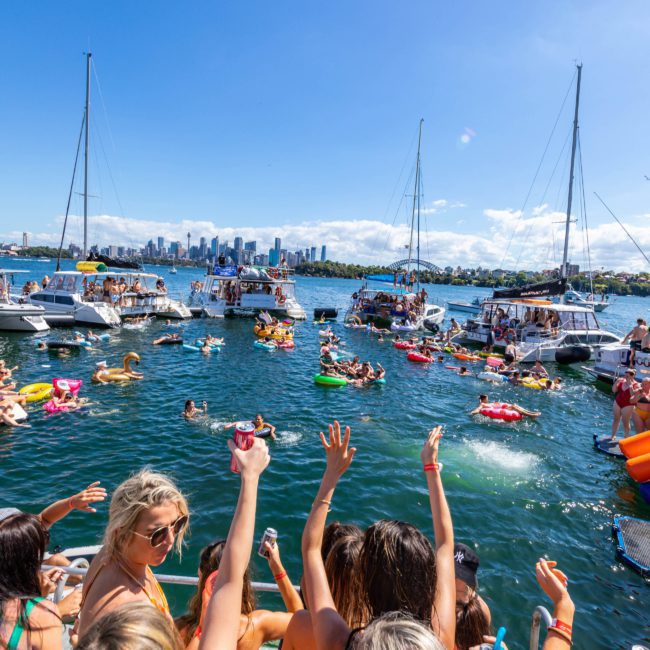 A group of people in swimsuits enjoys a Sydney boat party hire on a sunny day, with several boats and colorful floaties in the water near a city skyline.