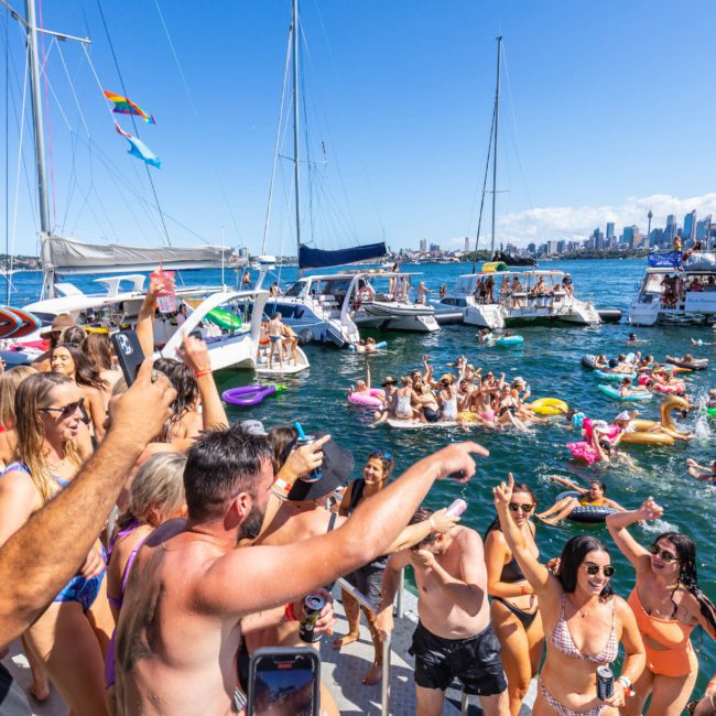 Large group of people in swimwear enjoying a lively Sydney boat party hire on a yacht-filled water, with many on inflatables, against a backdrop of city skyscrapers and a clear blue sky.