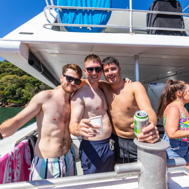 Three shirtless men stand together on a boat holding drinks, with one person taking a selfie. Other people are in the background enjoying the sunny day on what looks like a private yacht charter Sydney Harbour.
