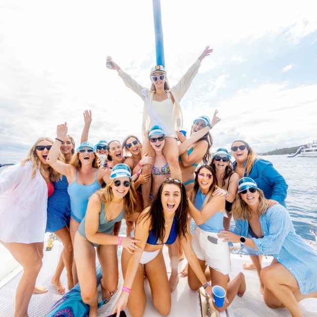 A group of women in casual and swimwear pose cheerfully on a private yacht charter in Sydney Harbour under a sunny sky, with a city skyline and other boats in the background.