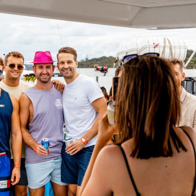 A group of six men posing for a photo on a boat, with one person taking the picture from behind. They are all holding beverages and casually dressed, with a scenic backdrop of water and land. Perfect for corporate boat events Sydney.