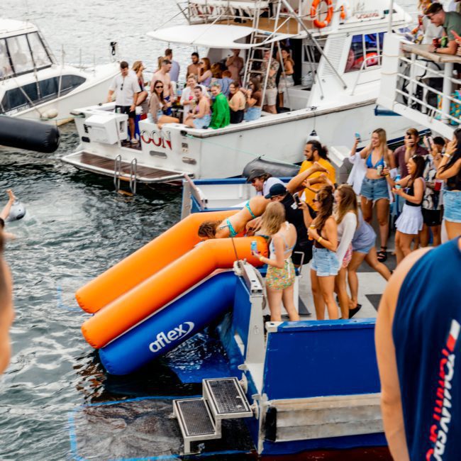 People are gathered on two boats, one of which has an inflatable water slide. Some are on the slide, while others stand nearby. The boats float on a body of water with hills in the background. Experience unforgettable moments with DJ boat hire Sydney for corporate boat events and more!