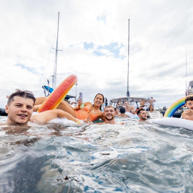 A group of people in water holding inflatable pool toys with boats in the background; some are smiling and engaging with the camera, enjoying a private yacht charter on Sydney Harbour.