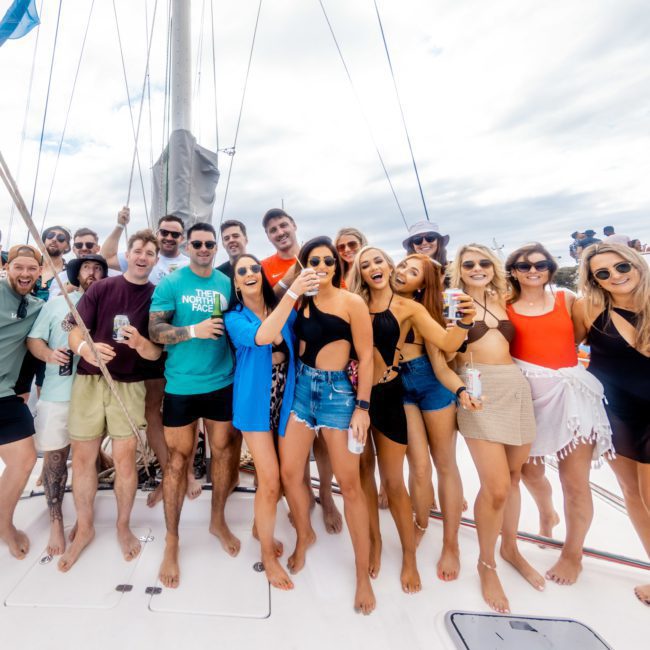 A group of people stand on a boat deck, smiling and holding drinks, enjoying their luxury yacht hire Sydney experience against a cloudy sky backdrop.