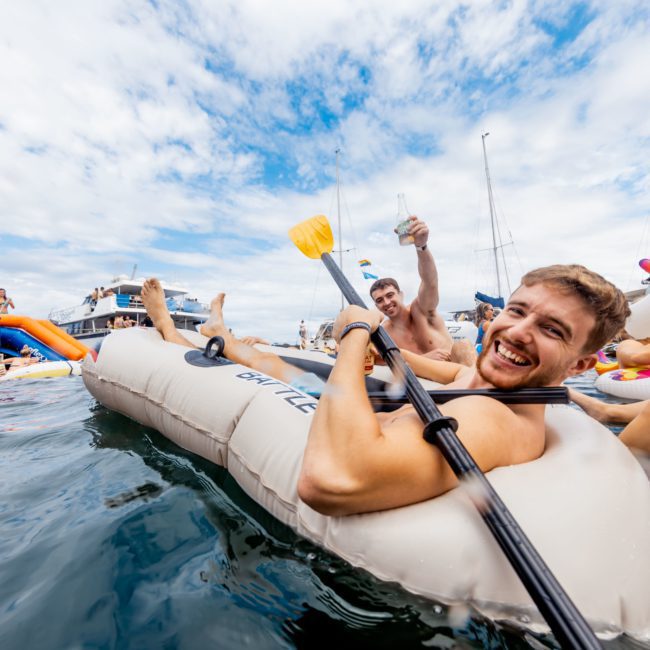 Two smiling men holding drinks are relaxing on inflatable rafts in the water, surrounded by boats and other people. One raft resembles a paddle boat, and the other is shaped like a unicorn. The scene unfolds near a luxury yacht hire in Sydney, adding a touch of elegance to their fun outing.