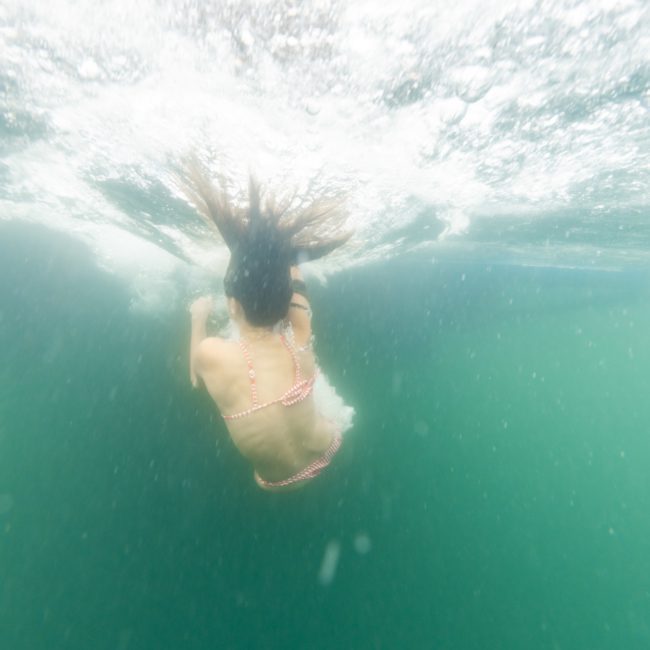 An underwater view of a person swimming in clear water, wearing a red and white striped swimsuit, beneath the pristine waves often seen during luxury yacht hire Sydney.