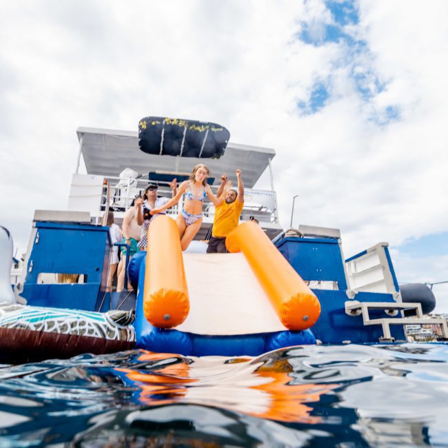 Three people descend an orange inflatable slide from a boat into the water, with one person pointing forward, during a lively corporate boat event in Sydney. The sky is partly cloudy.