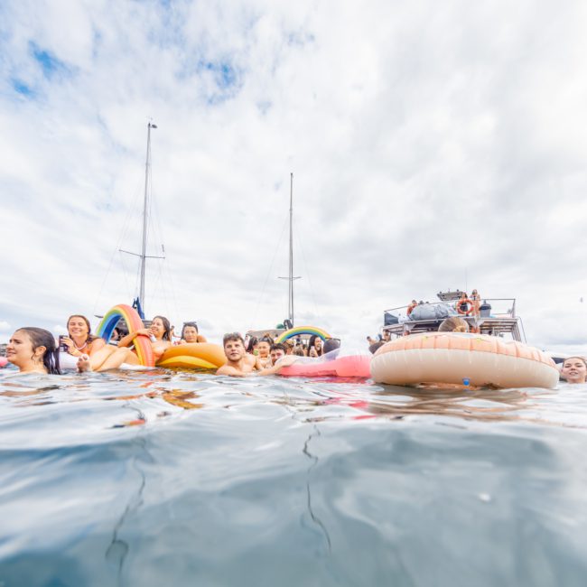 People in the ocean swimming and floating on inflatables, with boats and a cloudy sky in the background, enjoying a Sydney boat party hire.