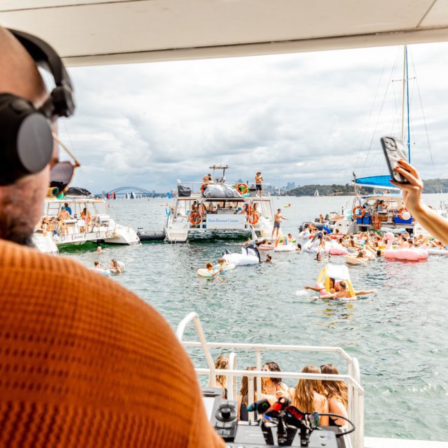 A DJ wearing headphones plays music on a boat during a lively Sydney boat party hire, while people on inflatables enjoy the water. Other boats and a cloudy sky are visible in the background.