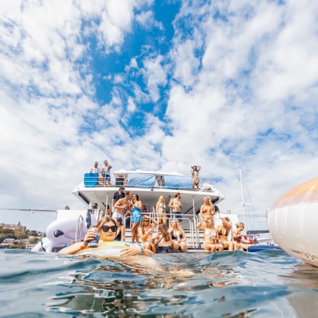 A group of people are gathered on a luxury yacht in Sydney, some holding drinks. The sky is partly cloudy.
