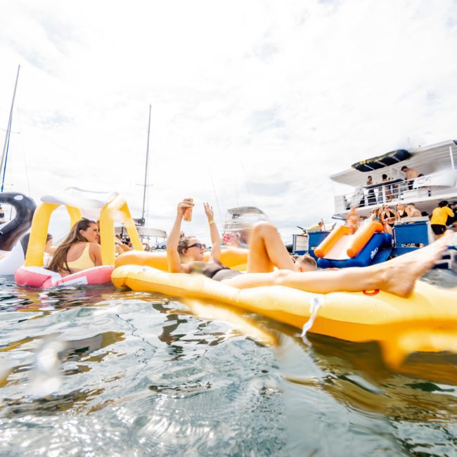 People are relaxing on inflatable floats in the water near a boat, enjoying a sunny day. Some are holding drinks and lounging as other boats, including luxury yacht hires in Sydney, are visible in the background.