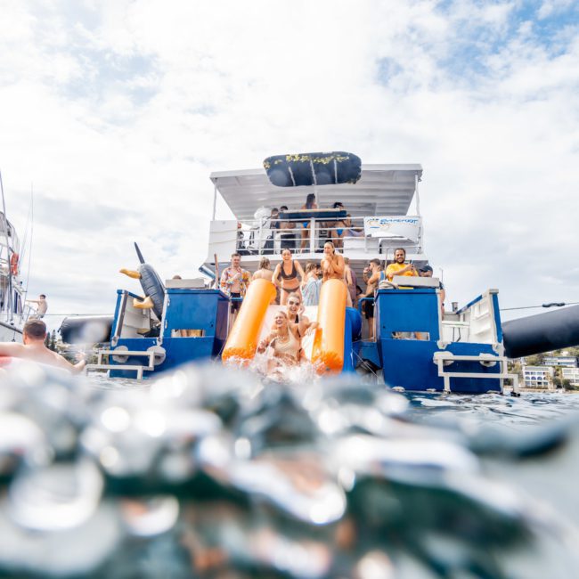 People are enjoying a day on the water near boats. Some are on the boat, while others swim nearby. The scene appears lively and recreational. It's reminiscent of a catamaran party in Sydney or perhaps even a corporate boat event happening right on these waters.