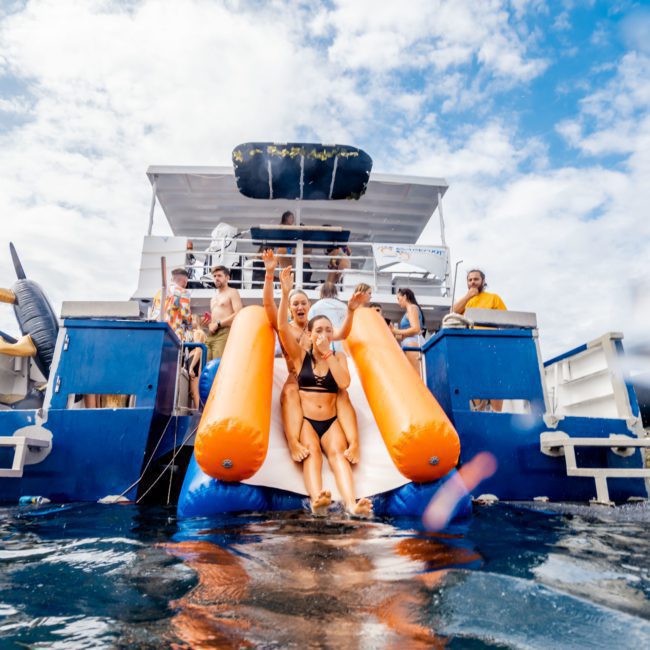 People slide down an inflated water slide attached to a double-decker boat surrounded by water, under a partly cloudy sky. Enjoy this thrilling attraction with Sydney boat party hire for an unforgettable day on the harbour.
