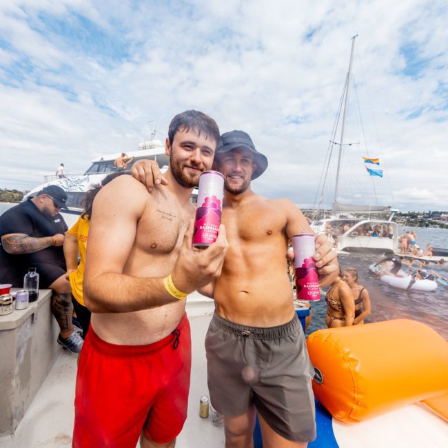 Two men stand on a boat holding beverage cans, smiling at the camera. Several people are in the background on other boats and inflatables, enjoying a sunny day on the water. Consider this for your next private yacht charter Sydney Harbour adventure!