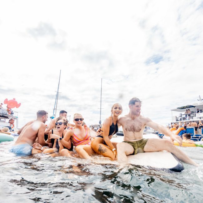 A group of people in swimsuits relax on a floating platform surrounded by boats and others in the water, enjoying a Sydney boat party hire experience.