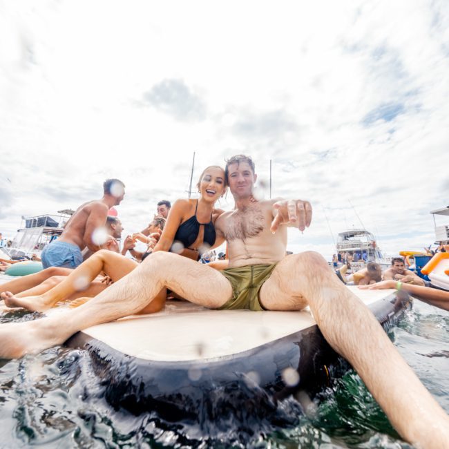 Two people sit on a paddleboard in the water, surrounded by others in a lively setting with boats in the background. It's a scene of fun and relaxation, perfect for those considering luxury yacht hire Sydney to elevate their day on the water.