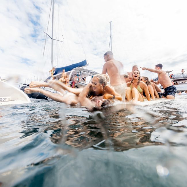 Group of people in swimsuits enjoying a playful activity in the water near anchored boats on a bright, sunny day during a private yacht charter in Sydney Harbour.