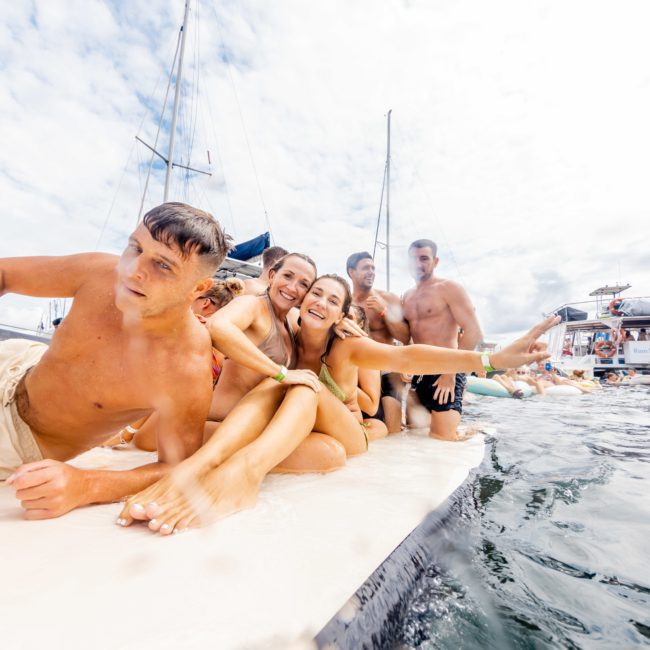 A group of people sits on a floating platform in the water, smiling and posing for the camera during a lively catamaran party in Sydney. Boats and other people are visible in the background, enhancing the festive atmosphere.