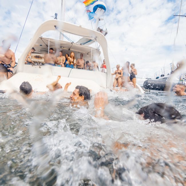 People are swimming and splashing around near a luxury yacht hire in Sydney, with many more individuals gathered on the boat and nearby vessels. The scene is lively and appears to be a social gathering on the water.