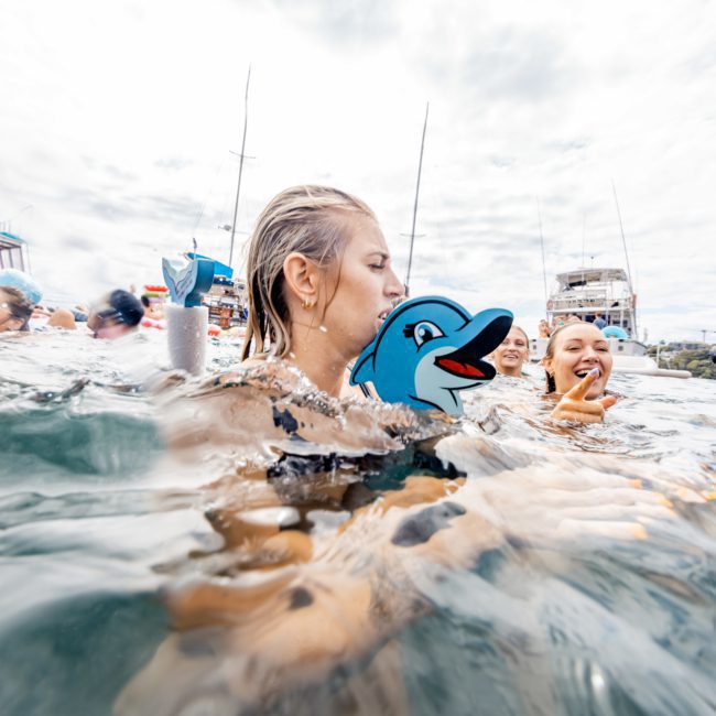 A group of people swim in the ocean near boats, with one person using an inflatable dolphin as a flotation device. Under a mostly cloudy sky, the scene could easily be part of a fun Sydney boat party hire.