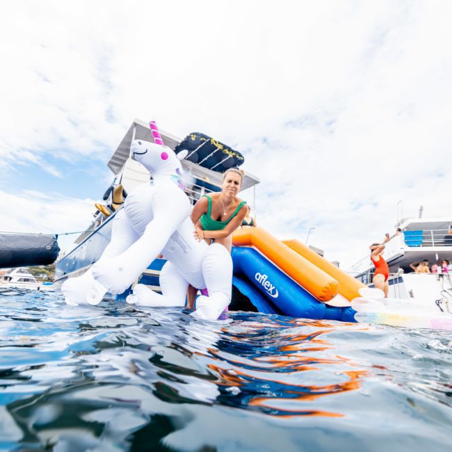 Description: Person sitting on an inflatable unicorn in the water near a boat, with several people in the background enjoying a private yacht charter on Sydney Harbour.