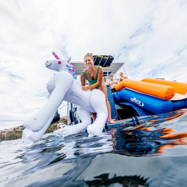 A person in a swimsuit relaxes on an inflatable unicorn float in the water near boats, enjoying the ambiance of a private yacht charter Sydney Harbour.