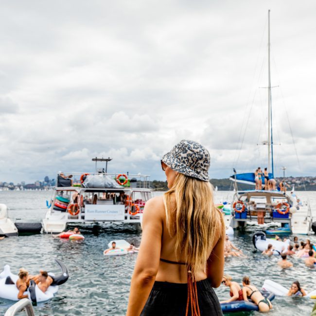 A woman with long hair and a bucket hat stands on a boat, overlooking a bustling group of people on inflatable rafts and boats under a cloudy sky. This vibrant scene is perfect for those considering Sydney boat party hire or even luxury yacht hire in Sydney for their next event.