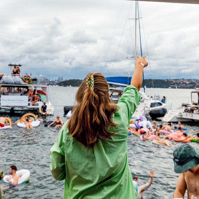 People in swimsuits enjoy a Catamaran party Sydney on the water with inflatables; boats are seen in the background. A woman wearing a green shirt is waving. The sky is cloudy.