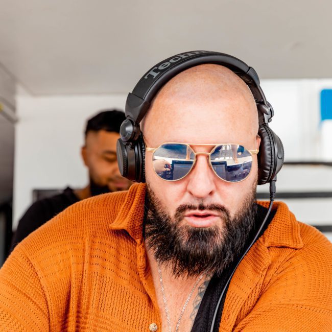 A bald man with a beard is wearing reflective sunglasses, headphones, and an orange shirt while appearing to DJ on a luxury yacht hire in Sydney. Another person stands in the background.