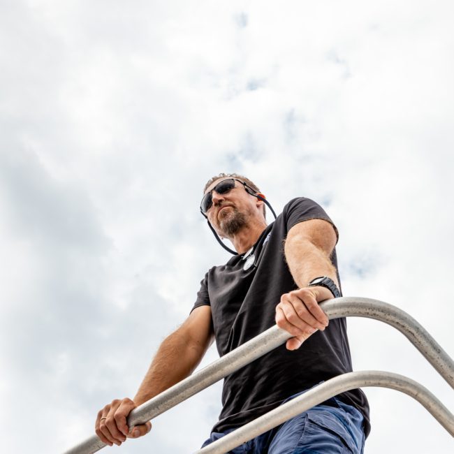 A man stands at a high vantage point overlooking the area while wearing sunglasses and a black shirt, with one hand on a metal railing. The sky is partly cloudy and in the distance, yachts dot Sydney Harbour.