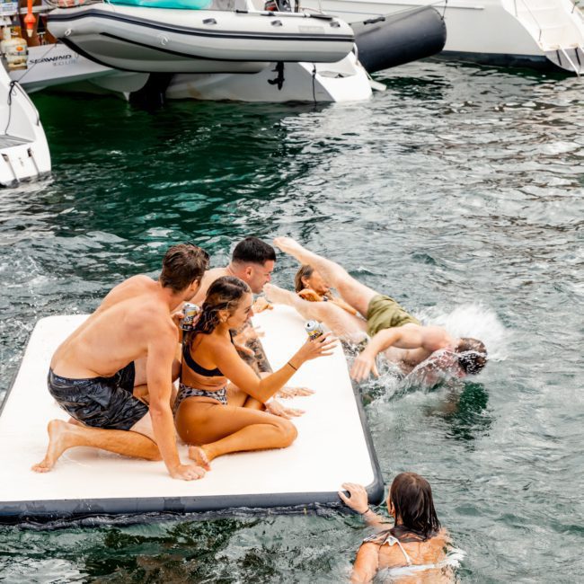 A group of people enjoying themselves on an inflatable floating platform next to boats on the water, with one person splashing into the water while others sit and observe during a lively catamaran party in Sydney.