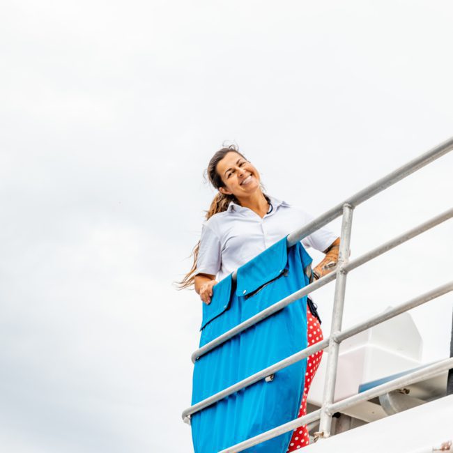 A woman is smiling while leaning on a railing on a boat, with the sky in the background. She is wearing a blue skirt and white blouse, enjoying the luxury yacht hire in Sydney.