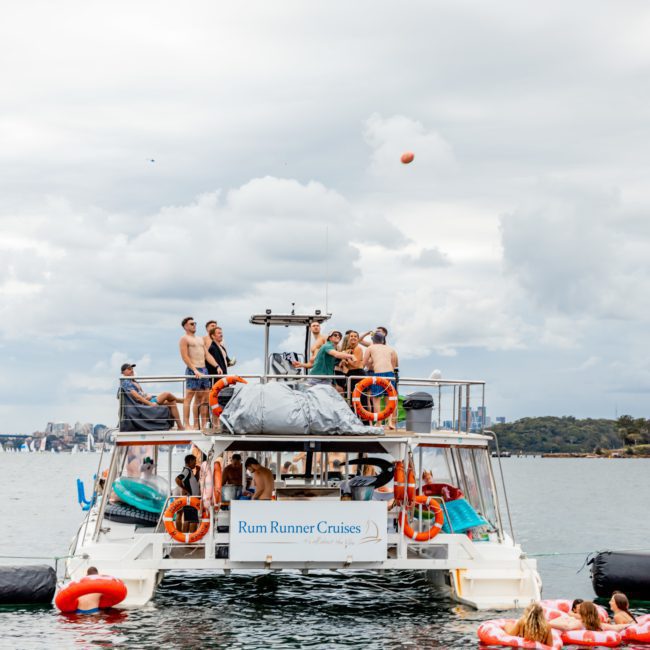 A group of people enjoys a Catamaran party Sydney on the stationary "Rum Runner Cruises" in a body of water, with several others floating nearby on inflatable rafts.