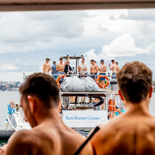 A group of shirtless people on the top deck of a boat named "Rum Runner Cruises" enjoys the water with a cloudy sky and distant land and structures in the background, showcasing an unforgettable Sydney boat party hire experience.