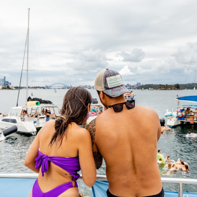 A man and a woman in swimwear stand on a boat, looking at a group of anchored boats with people swimming and socializing on a cloudy day. It's the perfect setting for a Sydney boat party hire experience.