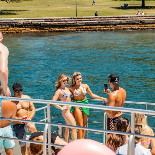 A group of people are gathered on a boat near the shore. Two women in the center pose for a photo while a man in a cap takes the picture. Others are seated or standing around them, enjoying their time on this private yacht charter Sydney Harbour.