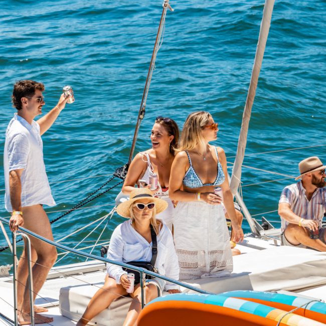 A group of people relaxes and enjoys beverages on a luxury yacht in Sydney. One person stands and raises a drink while others sit or lean on the boat's railing. A paddleboard labeled "SPINERA" is attached to the boat, highlighting the opulent experience of Luxury yacht hire Sydney.