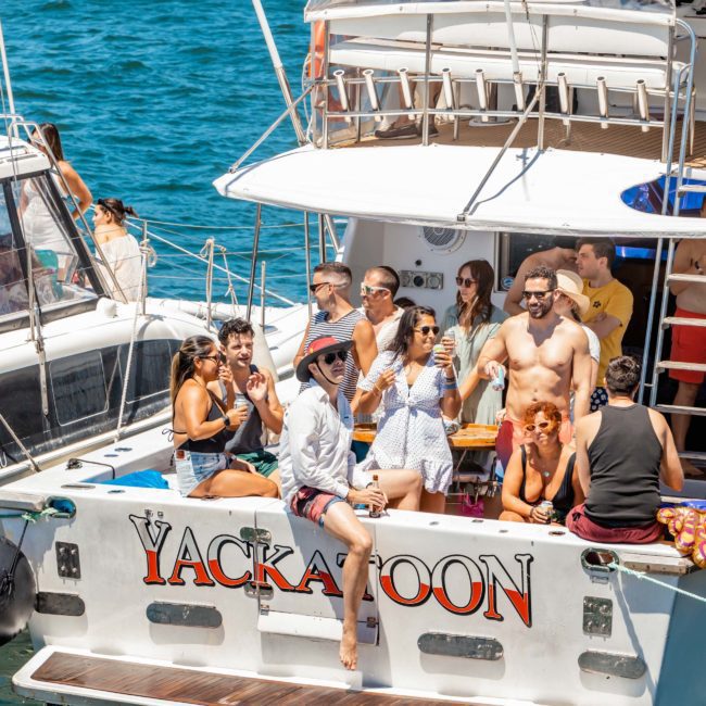 A group of people socialize and relax on the deck of a catamaran named "Yackatoon" on a sunny day. The boat is docked near a body of water with clear blue skies above, setting the perfect scene for a Sydney boat party hire.