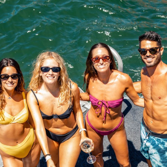 Four people in swimwear standing on a boat, smiling and holding drinks, with water visible in the background. Enjoy the vibrant atmosphere of a DJ boat hire Sydney experience.