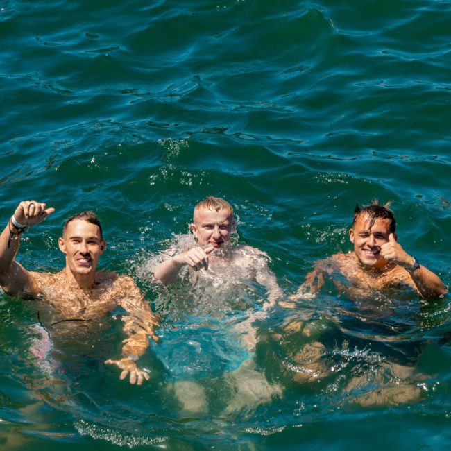 Three men swimming together in deep blue water, smiling and waving at the camera during a Catamaran party Sydney.