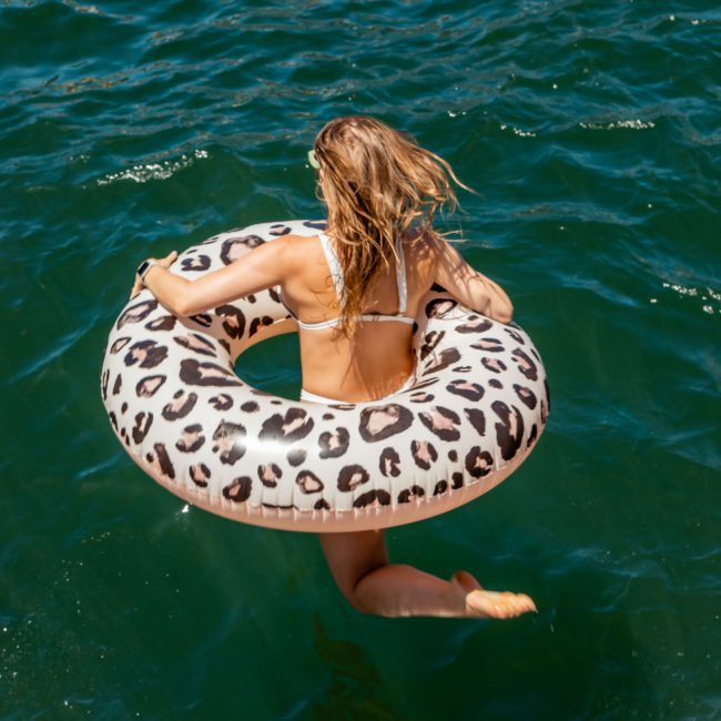 A woman holding a leopard-print inflatable ring jumps into a body of water while a man stands on a metal ladder beside her, reminiscent of the fun and excitement of a DJ boat hire Sydney adventure.