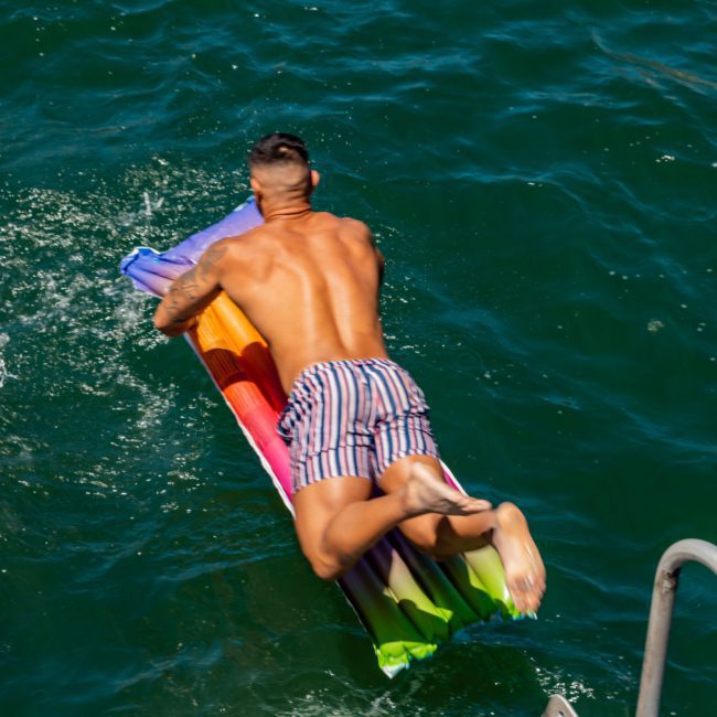 A man in striped shorts dives onto a colorful inflatable pool raft in a body of water, near a luxury yacht hire in Sydney.