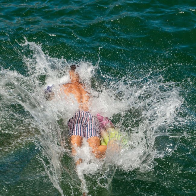 Two people mid-air diving into water, creating a big splash, with a metal ladder visible in the foreground during a DJ boat hire Sydney event.