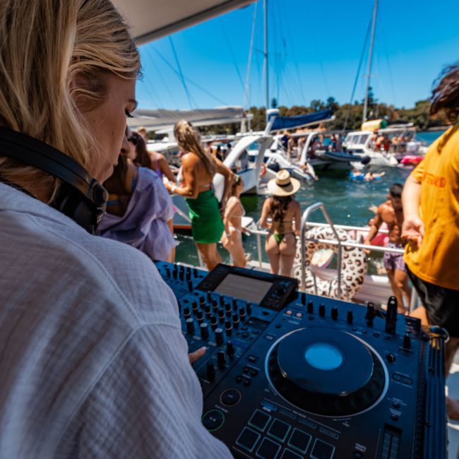 A DJ in white clothing is playing music on a boat deck in front of a lively crowd. Several other boats can be seen in the background on a sunny day. Experience this vibrant atmosphere with DJ boat hire Sydney for your next event.