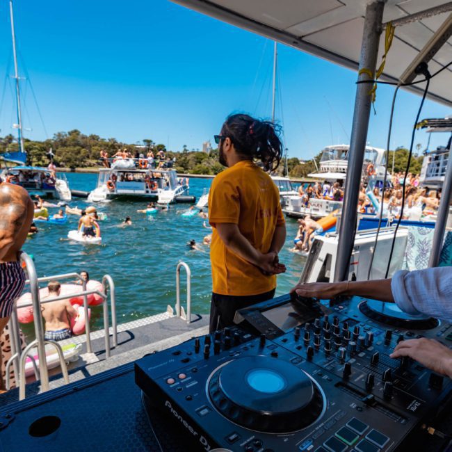 People are enjoying a Sydney boat party hire on a catamaran docked near others in a sunny waterside area. A DJ is playing music, and several people are swimming and relaxing on floaties nearby.