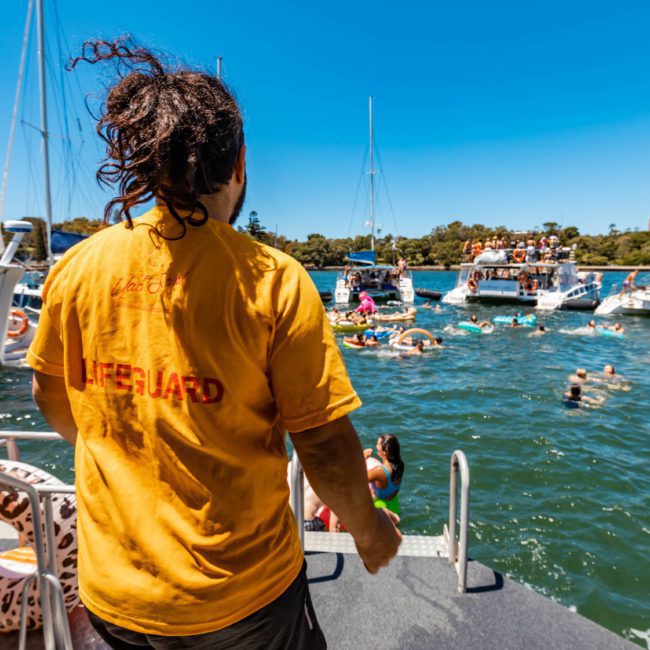 A lifeguard in a yellow shirt overlooks a crowded, sunny marina where people swim and float near various boats. A nearby catamaran party adds to the lively atmosphere under the clear blue sky.