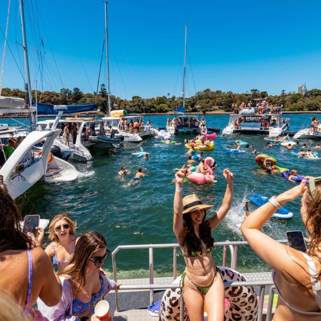 People are gathered on boats and inflatables in a sunny waterway, enjoying a festive atmosphere. Participants are swimming, dancing, and relaxing under a clear blue sky. A luxury yacht hire Sydney adds an extra touch of elegance to the scene.
