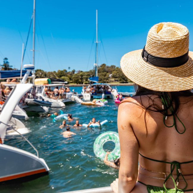 A woman wearing a straw hat views a bustling scene of people swimming and boating on a sunny day, with private yacht charters in Sydney Harbour adding an air of sophistication.